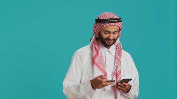 Smiling adult using card to pay online while shopping on internet store website with smartphone. Middle eastern guy in islamic clothing making baking transfer to buy something on sale. photo