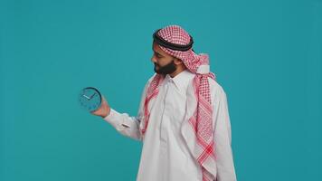 Middle eastern man checks wall clock to look at time, using timepiece to see hour and minutes. Young adult in national muslim clothing examining local timezone, standing over studio background. photo