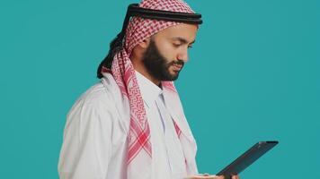Middle eastern adult browsing device in studio, navigating websites online on digital gadget. Muslim person in traditional clothes using tablet to check internet pages, texting on social media. photo