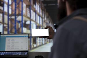 Storehouse employee looking at phone with blank display template, verifying industrial stock labels and order receipts. Supervisor providing cargo merchandise for supply chain contracts. photo