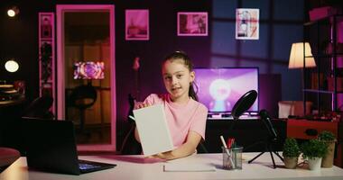 Cute young media star assessing book received as homework from school after reading it, expressing satisfaction with plot. Kid content creator shooting lecture review for online channel photo
