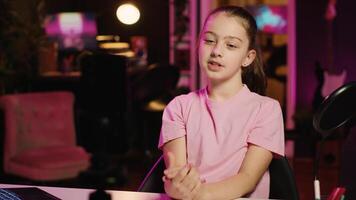 Charming young girl shooting for social media platform in living room, talking about her day. Kid entertaining followers, doing content creation in neon lit apartment using smartphone on tripod photo