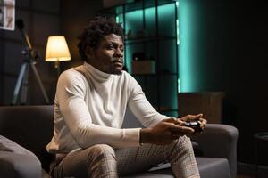 BIPOC player at home gaming and enjoying leisure time. Man sitting on couch in cyan neon illuminated living room, playing videogame on console, relaxing after hard day at work photo