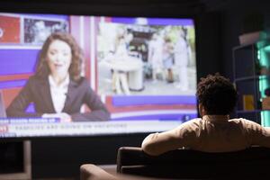 Man sitting on couch in front of ultrawide smart TV displaying news program broadcasting. Person watching big television screen showing VOD channel with newscast coverage photo