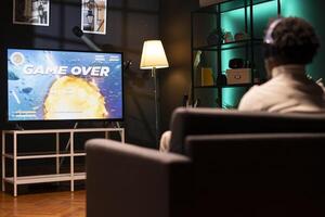 Gamer wearing headphones frustrated by seeing game over message on smart TV while playing arcade space shooter videogame. Man in home theatre spending time on gaming console, disappointed after losing photo