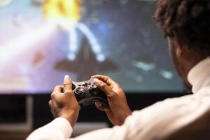 Controller held by gamer sitting in front of large projector displaying videogame. Close up shot of gamepad used by man enjoying gaming session on console connected to ultrawide smart TV photo