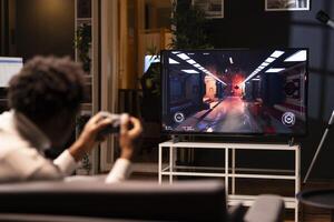 Gamer using controller to play first person shooter videogame on smart TV, shooting robotic enemies. African american man in home theatre using high tech gaming console gamepad photo