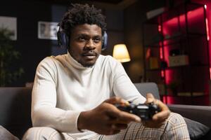 BIPOC man playing videogames using controller, having fun in red neon lit apartment. Gamer competing in online multiplayer game on gaming console, using headphones to talk with teammates photo