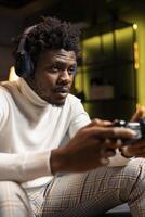 African american man playing videogame in yellow neon lit apartment, holding controller. Gamer participating in PvP online multiplayer game using console system and headphones for immersive experience photo