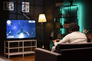 BIPOC man in home theatre using cloud gaming service to play demanding science fiction videogame on smart TV. Gamer enjoying high quality game, streaming it online, flying spaceship with controller photo