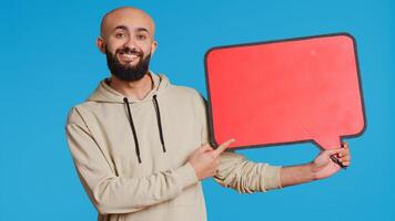 Arab person holding red speech bubble icon in studio, creating web advertisement for new product commercial. Middle eastern man shows isolated cardboard sign for ad. Camera 2. Handheld shot. photo