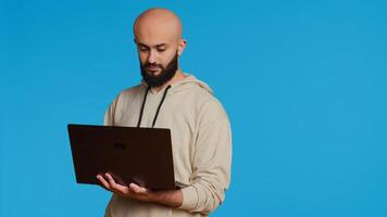 Middle eastern man works with cyber security on laptop, trying to do some software programming and report bugs. Young adult operating as a system developer in studio. Camera 2. Handheld shot. photo