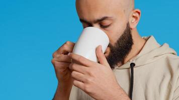 Middle eastern guy drinking cup of coffee in studio, enjoying fresh aroma of caffeine refreshment. Arab person standing over blue background, taking a sip from mug. Camera 2. Handheld shot. photo