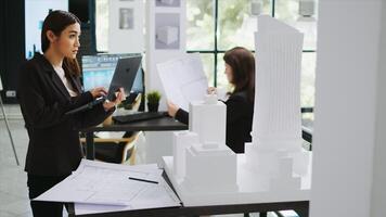 Asian contractor takes notes about 3d printed model on laptop, examining outline and scale of building maquette in architectural agency office. Cad specialist measuring layout for new project. photo