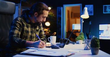 Self employed broker investor in home office checking stock exchange profit, calculating using pen and paper. Freelancer in apartment looking at market shares growth, writing on notepad, camera B photo