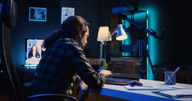 Experienced programmer writes code on laptop screen while in apartment office using Java programming languages. Developer remotely working on fixing database errors while working from home, camera B photo