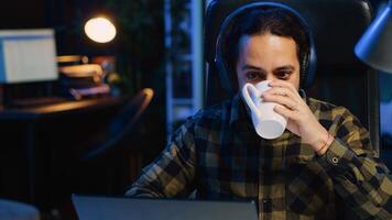 Happy man drinking coffee and writing emails at home office desk while listening music. Smiling remote worker doing b2b networking over internet, wearing headphones, enjoying hot beverage, camera A photo
