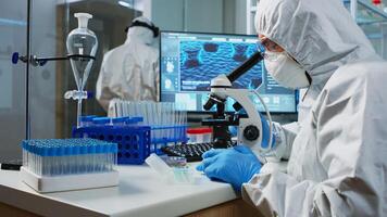 Scientist in protection suit looking through a microscope in equipped lab. Team of chemists examining virus evolution using high tech for scientific research of vaccine development against covid19. photo