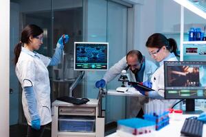 Scientist analyzing blood sample in vacutainer with researching team looking through microscope. Chemists in pharmaceutical lab examining sample for medical experiment with technology for medicine industry. photo