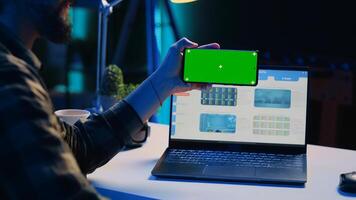 Man sitting on chair at home office desk in neon illuminated apartment holding green screen phone while working. Person in living room looking at business graphs on laptop and at mockup smartphone photo