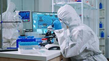 Chemist in protection suit typing on pc and analyzing virus sample using microscope in scientific lab. Team of doctors examining vaccine evolution using high tech researching diagnosis against covid19 photo