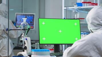 Man researcher in coverall looking at chroma key computer in modern equipped lab. Team of microbiologists doing vaccine research writing on device with green screen, isolated, mockup display. photo
