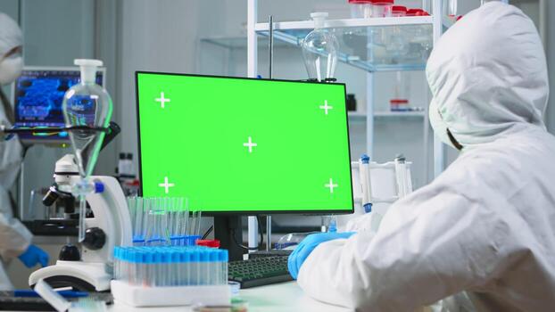 Scientist man wearing protection suit typing on computer with green mockup in modern equipped lab. Team of microbiologists doing vaccine research writing on device with chroma key, isolated display. photo