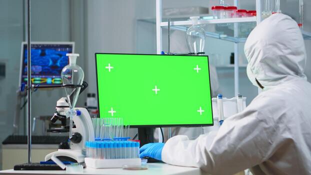 Doctor with coverall working at computer with green screen in modern equipped lab. Team of microbiologists doing vaccine research writing on device with chroma key, isolated, mockup display. photo