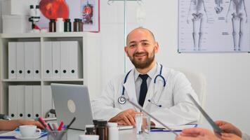 Doctor in lab coat and stethoscope over neck typing on laptop at desk looking at camera and smiling in busy workday in medical office. Specialist writing on notepad during brainstorming in meeting room photo