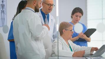 Team of doctors standing in conference hospital room, senior doctor discussing about treatment for patient looking in laptop. Coworkers in white coats working together analysing symptoms of disease photo