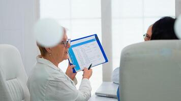 Elderly physician is brainstorming to discuss medical issues pointing on clipboard presenting list of sick patients. Team of doctors working in hospital office discussing symptoms of disease photo