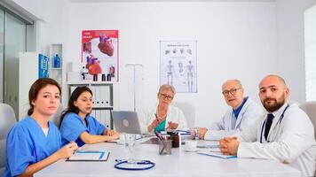 Group of specialist doctors having meeting, discussing on webcam sitting at desk in medical conference hospital room using modern technology. Professional team looking at camera and talking photo