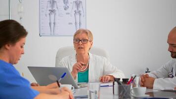 Senior surgeon explaining treatment to coworkers presenting plan during medical conference sitting at meeting desk in modern clinic. Profesional team taking notes while specialist doctor discussing. photo