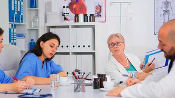 Senior specialist doctor is brainstorming to analyze medical problems pointing on clipboard showing medical statistics. Team of doctors working in hospital office discussing symptoms of disease photo