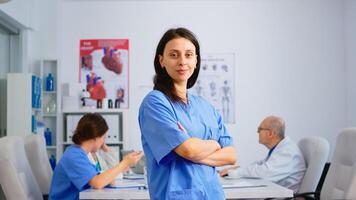Nurse standing in front of camera smiling in medical conference meeting office. Team of specialist doctors working in background talking about symptoms of disease in clinic room photo