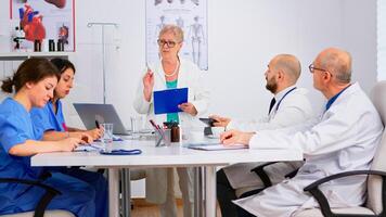 Portrait of mature woman doctor heading committee meeting at medical council presenting new medical procedures standing at desk. Group of physicians talking about symptoms of disease in clinic office photo