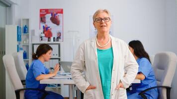 Portrait of elderly doctor standing in front of camera smiling in medical conference meeting office. Team of specialist doctors working in background talking about symptoms of disease in clinic room photo
