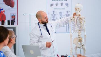 Specialist doctor in white coat showing to colleagues body bone functionson using skeleton model. Medic pointing and explaining to coworkers medical procedures sitting in hospital meeting room. photo