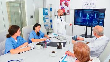 Mature medical physician explaining treatment to nurses during healthcare seminar pointing at digital monitor. Clinic herapist discussing with colleagues about disease, medicine professional. photo