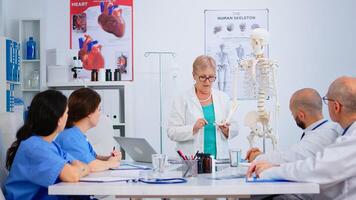 Elderly woman doctor showing the work of human's hand on skeleton anatomical model. Specialist medic explaining diagnosis to colleagues standing in front of desk in hospital meeting room. photo
