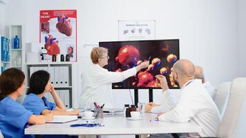 Senior doctor pointing on monitor in hospital office explaining virus development using digital screen in front of coworkers during brainstorming. Team of physicians analysing diagnosis of patients photo