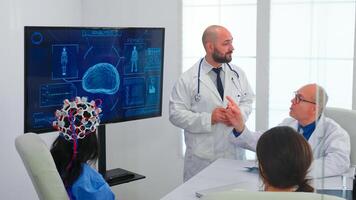Doctor talking about brain activity during conference with medical staff and nurse wearing headset with sensors. Monitor shows modern brain study while team of scientist adjusts the device. photo