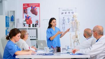 Doctor describing the layout of the human spine in front of colleagues using skeleton model explaining medical procedures standing in hospital office. Physicians discussing about symptoms of disease photo