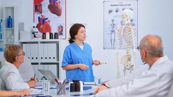 Medical young nurse presenting anatomy with human skeleton model describing the layout of the human spine. Explaining diagnosis to colleagues standing in front of desk in hospital meeting room. photo