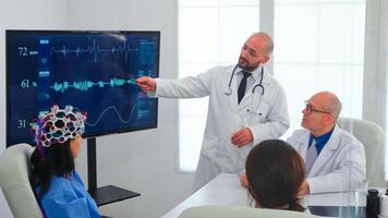 Nurse wearing headset with sensors during neuoroscience experiment in medical conference team. Monitor shows modern brain study while team of scientist adjusts the device working in hospital boardroom photo