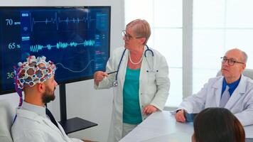 Scientist doing brain analysis using headset with sensors on doctor in hospital meeting room. Monitor shows modern brain study while team of scientist adjusts the device working in conference office photo