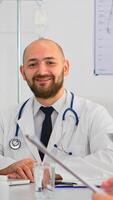 Doctor in lab coat and stethoscope over neck typing on laptop at desk looking at camera and smiling in busy workday in medical office. Specialist writing on notepad during brainstorming in meeting room photo