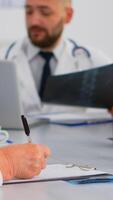 Close up of senior woman doctor taking notes on clipboard while radiologist coworkers discussing in background analysing x-ray and writing on laptop. Profesional teamworker having medical meeting photo