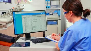 Orthodontist nurse looking at list with appointments in computer and taking notes on clipboard while dentistry doctor is working with patient background examining teeth problems.. photo