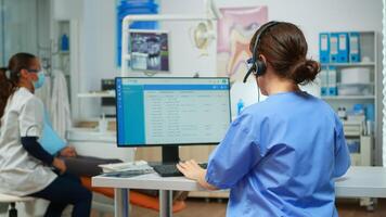 Dentist assistant making appointments using headset sitting in front on computer while doctor is working with patient in background examining teeth problem. Nurse taking notes in stomatological office photo